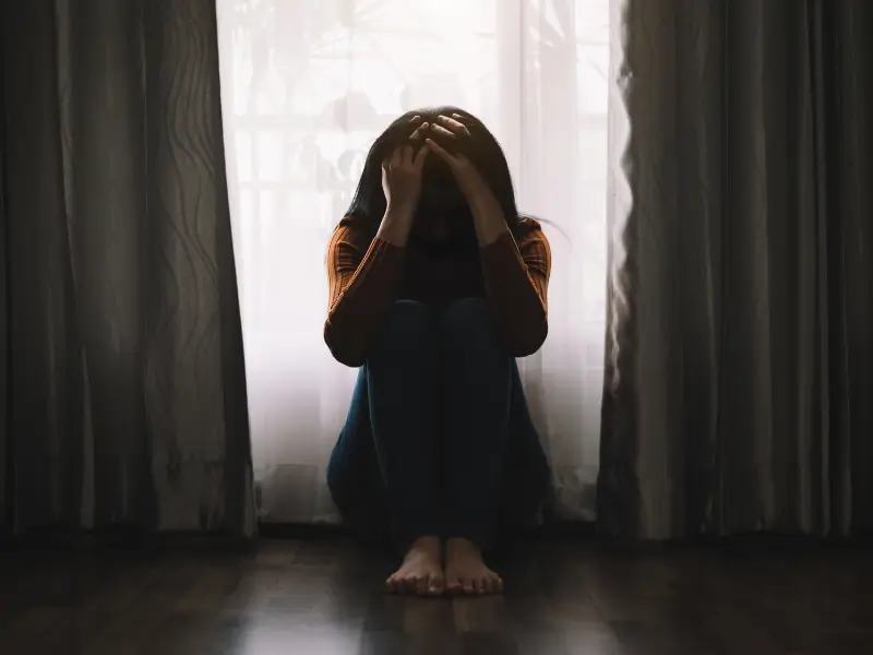 A woman sitting in the dark, against a window hidden by curtains. She is on the floor, holding her head with her two hand, hiding her face, looking depressed.