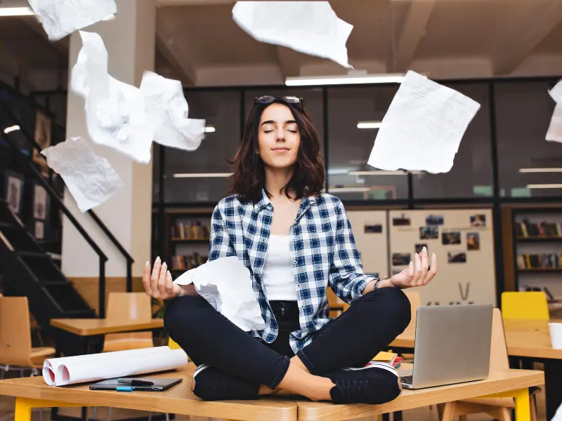 A young woman sitting on a table in a corporate office kitchen. She is sitting in a yoga pose with her eyes closed and looking relaxed. In the meantime, papers are flying around her.