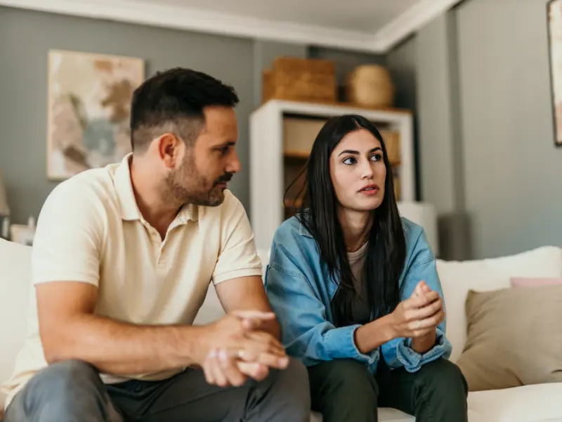 A man and a woman, a couple, sitting on a couch, close to each other but not touching, looking anxious and talking to someone in front of them.