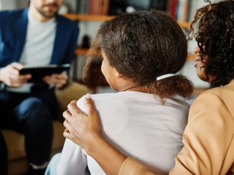 A woman and her daughter talking to a therapist. We cannot see their faces but the mom is looking at her daughter with her hand on her daughter's shoulder, showing support.