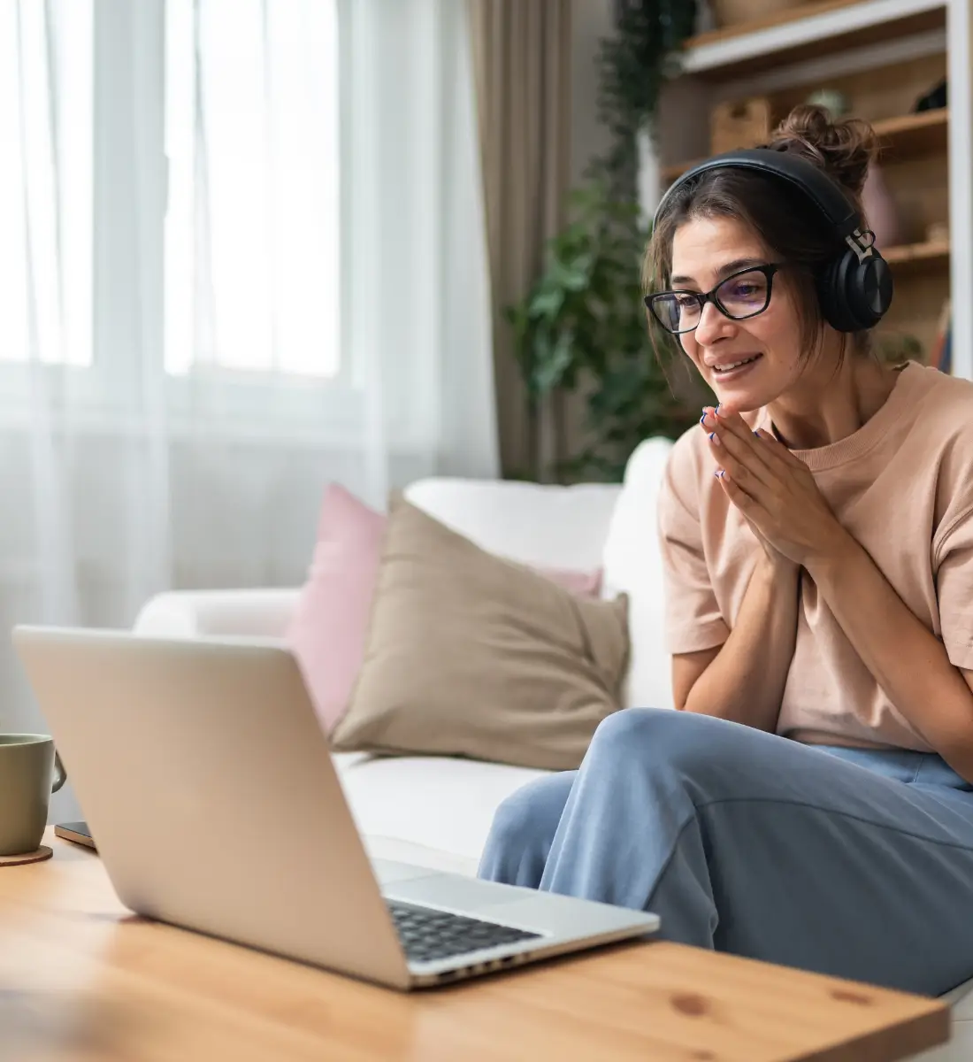 A young woman sitting on her couch, with headphones on, facing a laptop. She is smiling and talking to her therapist online.