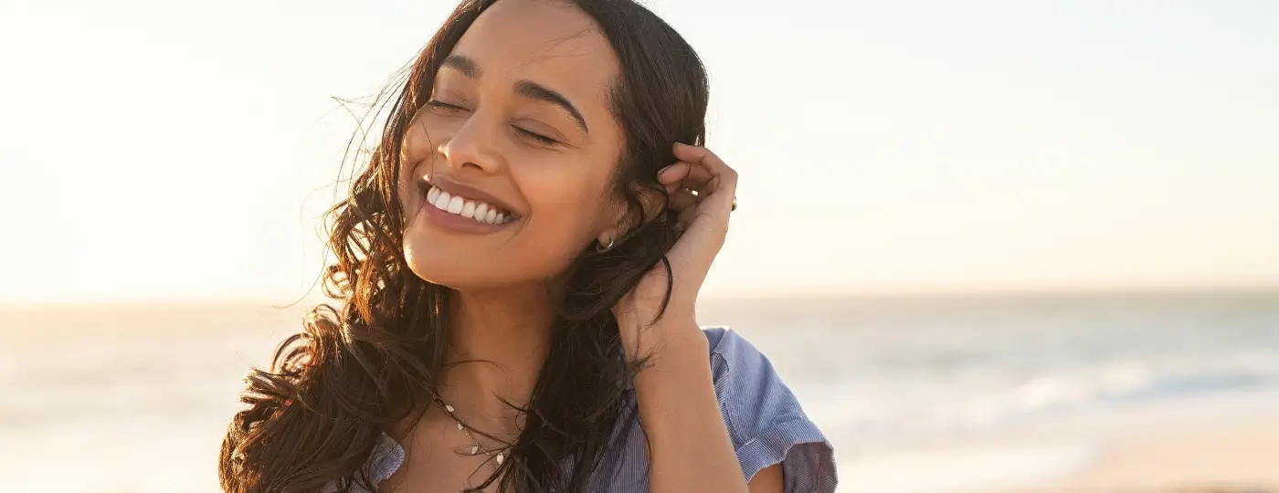 A young woman on a beach. We can only see her face, she is looking happy and smiling with her eyes closed.