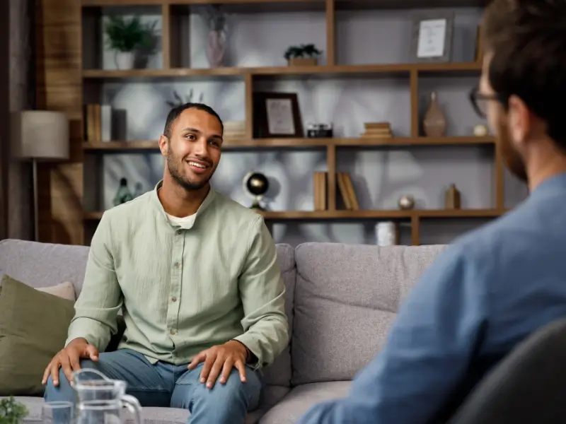 A young man sitting on a couch, talking to a therapist, smiling.