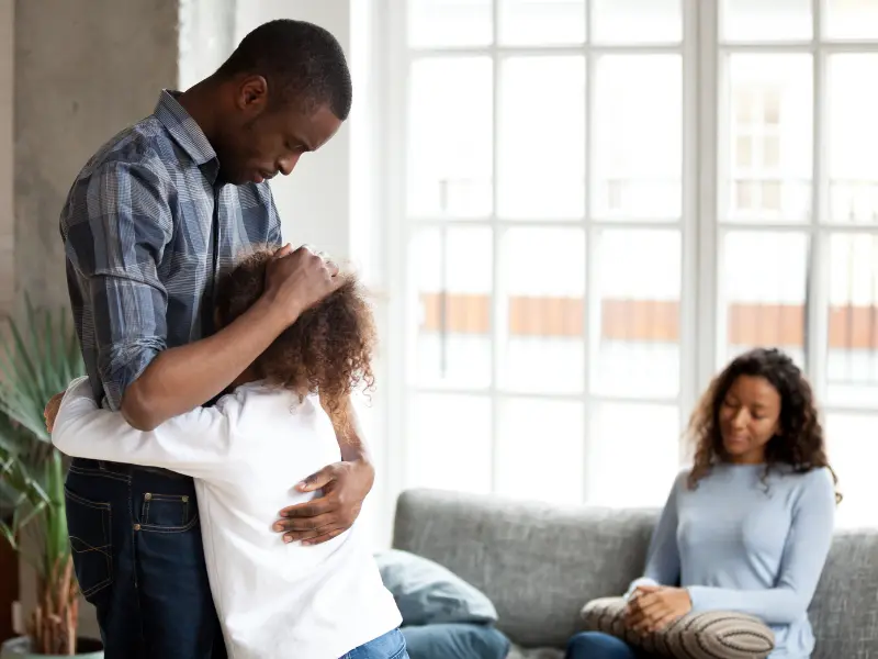 A man standing and hugging his daughter. The mom is sitting on the couch in the background, looking sad.