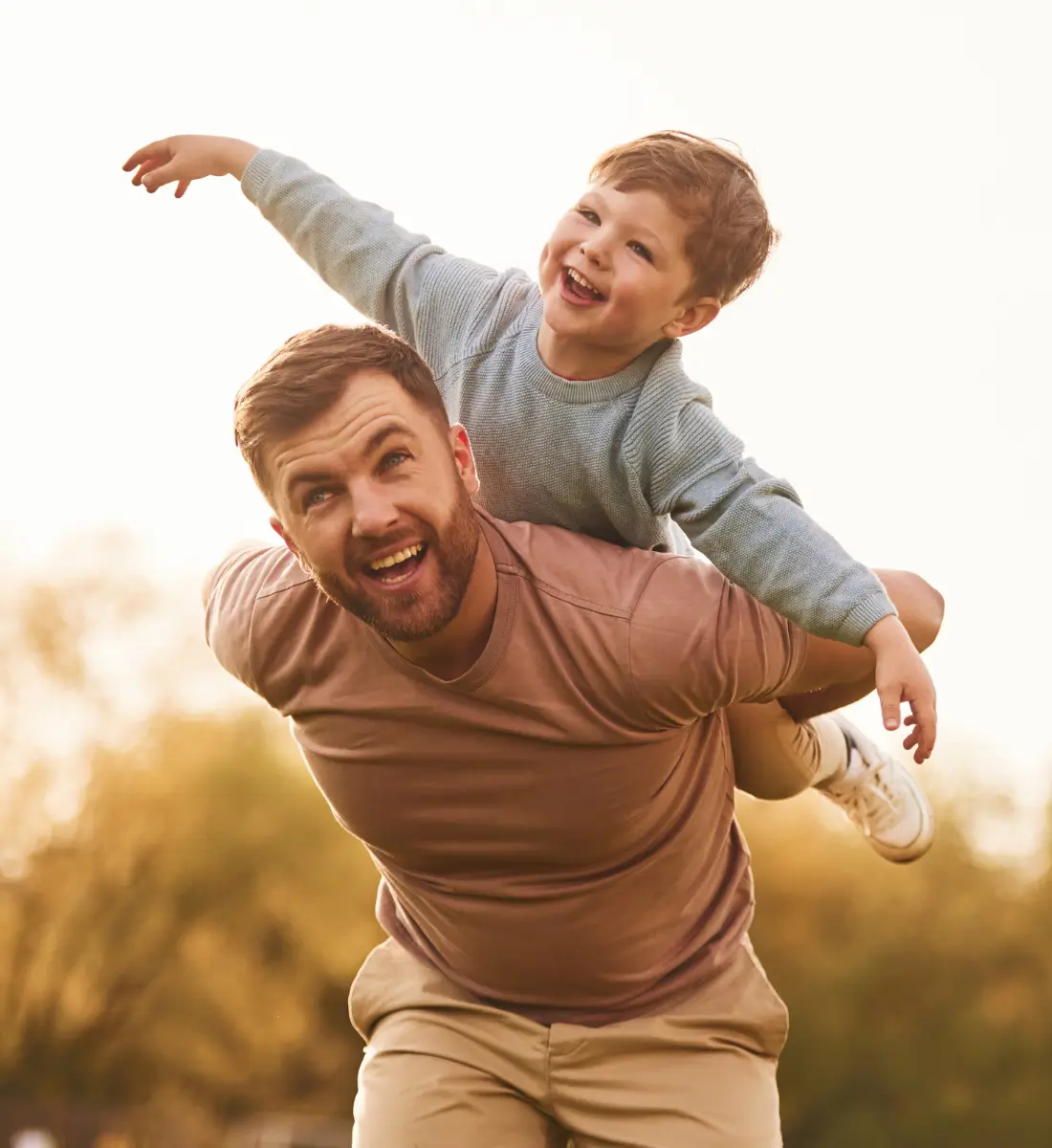 A dad playing with his son outside. The son is on the dad's back, with his arms wide like an airplane. Both are looking happy and having fun.