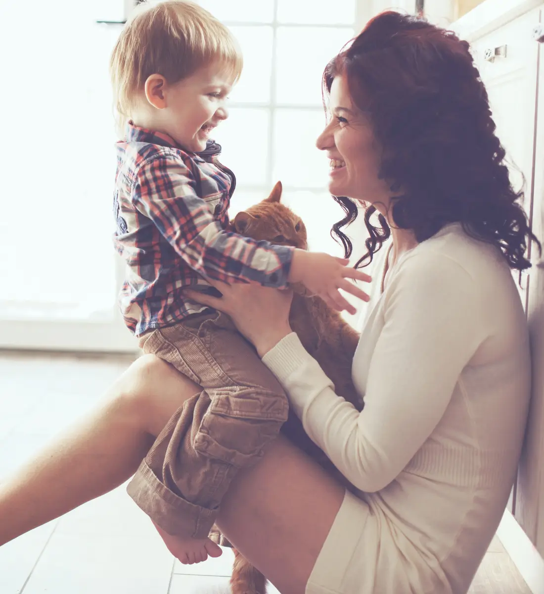 A woman sitting on the floor with her son sitting on her knees, facing her. Both are looking at each other and laughing.