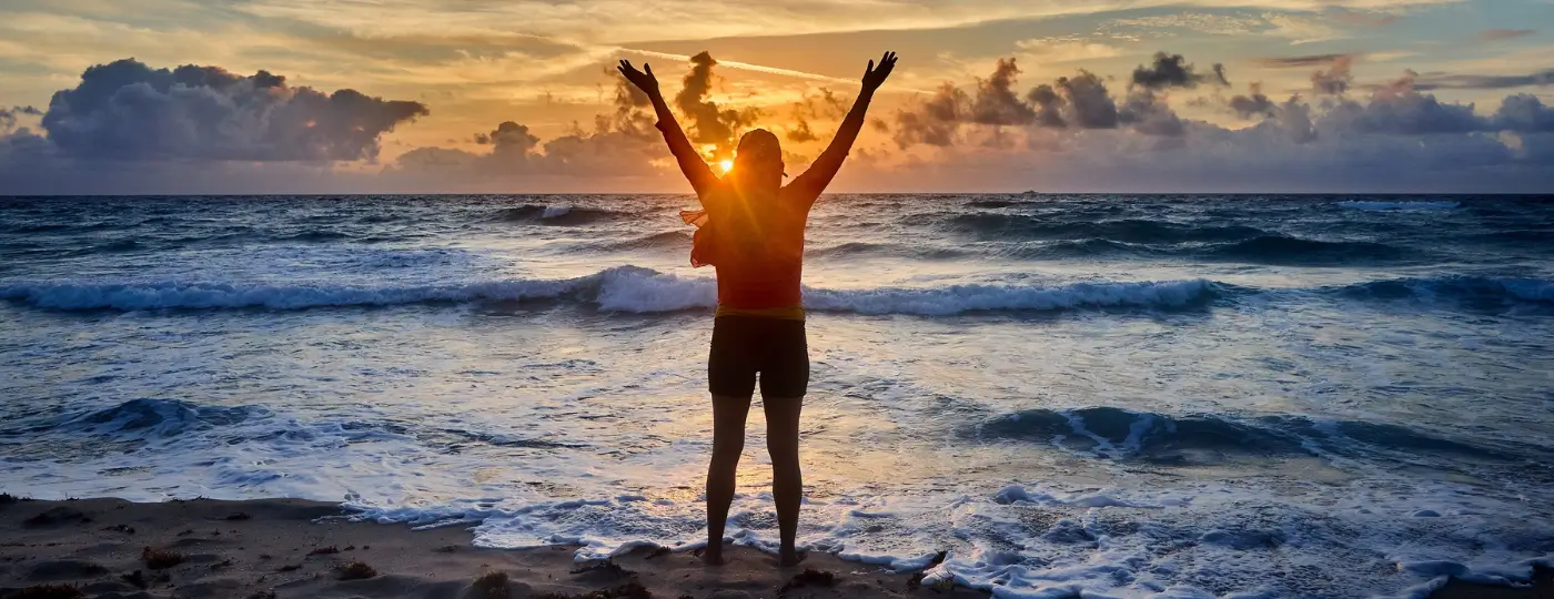 A person facing the ocean at sunset, raising their arms in a freedom posture