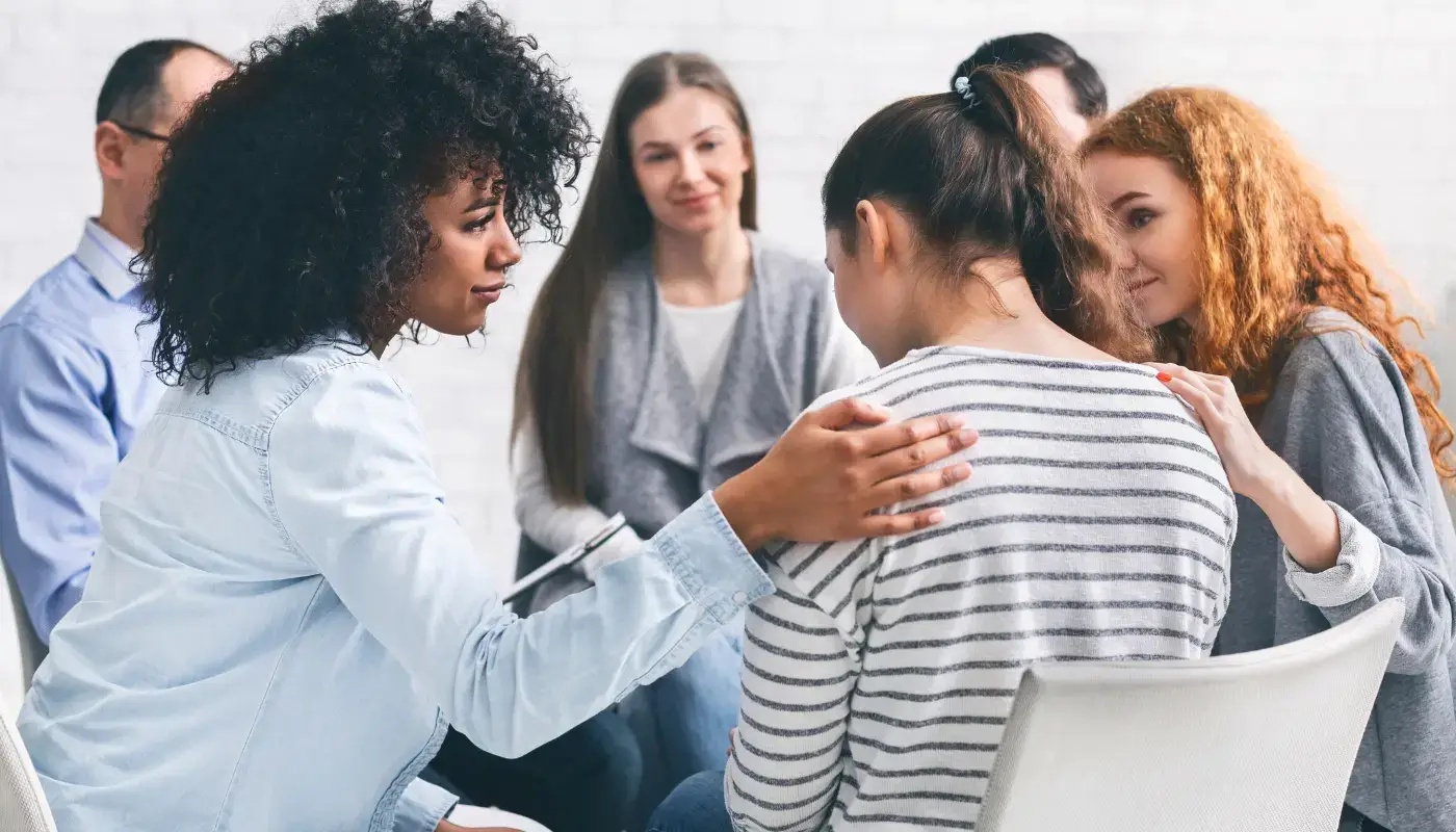 A group of people, sitting in circle in an addiction or alcoholic anonymous. A few of them are showing support to a woman, putting their hands on her shoulders and looking compassionate