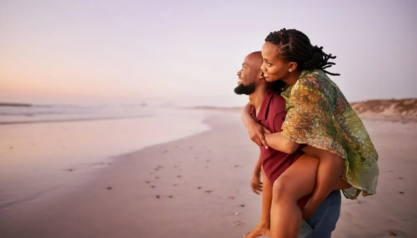 A couple embracing each other on the beach, the woman is on the man's shoulders, looking at the ocean at sunset