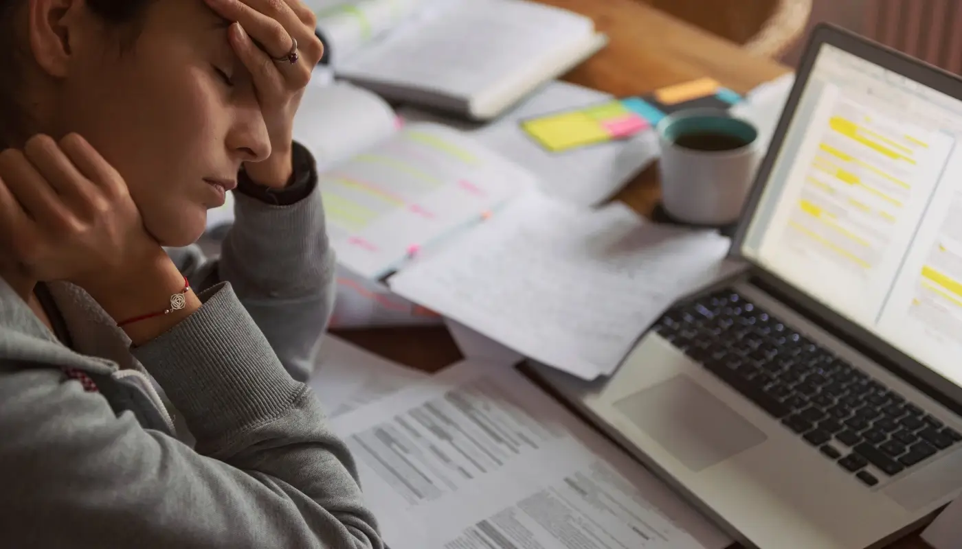 A person in front of their computer and books, looking overwhelmed, taking their head into their hands