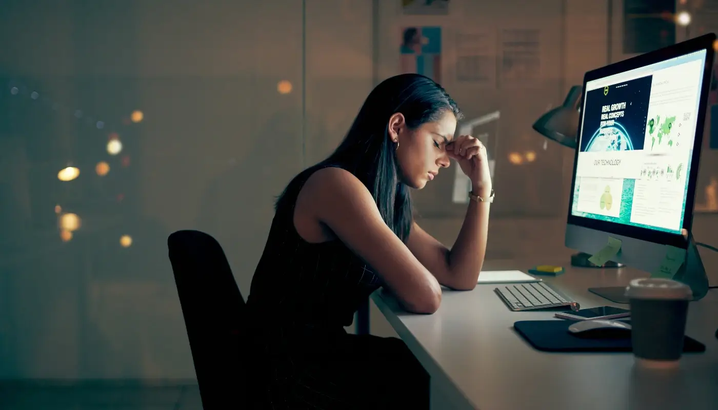 A woman sitting down at her desk, in front of her computer. She is closing her eyes with her hand on her forehead, looking stressed and overwhelmed.
